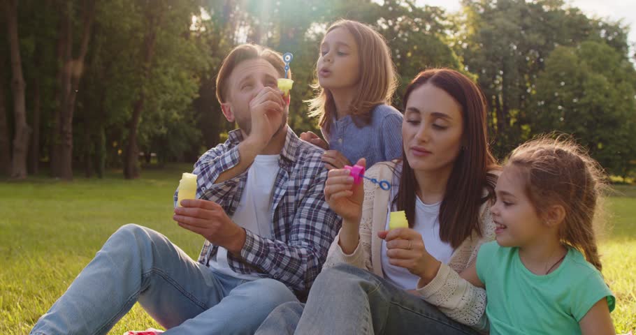 Happy young family sitting on grass in sunny park and blowing soap bubbles together. Smiling parents and children having fun during outdoor activity while playing and enjoying leisure time in nature.