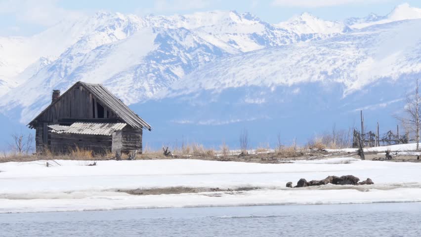 An old wooden fishing hut on the shore of Lake Baikal against the backdrop of snow-capped mountains in early spring.