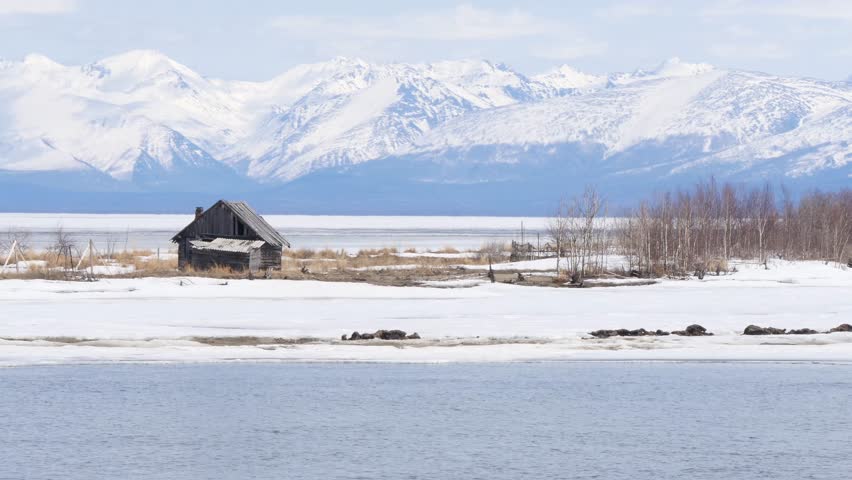 An old wooden fishing hut on the shore of Lake Baikal against the backdrop of snow-capped mountains in early spring.