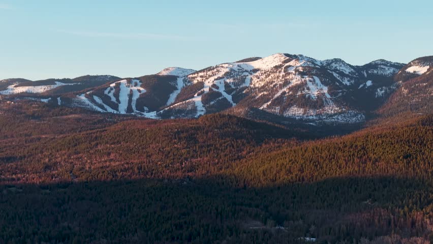 Aerial view of snow-capped mountain range and evergreen forest near Whitefish, Montana, captured at sunrise with golden light.