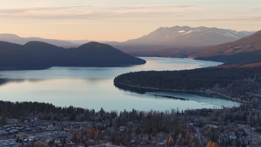 Aerial drone footage shows a winding lake bordered by evergreen forest and distant snow-capped mountains near Whitefish Montana at sunrise No animals are visible
