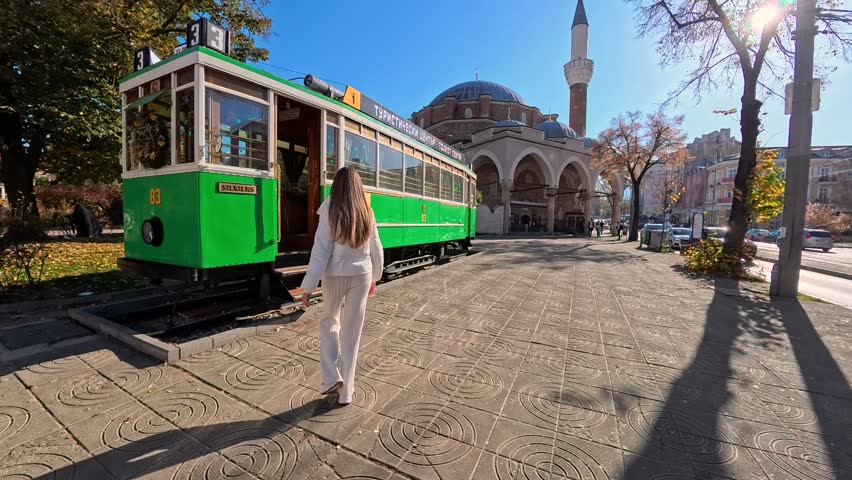 Sofia, Bulgaria - 11.02.2025. Happy female tourist posing near vintage tram in Sofia city center, Bulgaria, smiling traveler in white outfit enjoying sunny day, urban travel lifestyle, sightseeing and