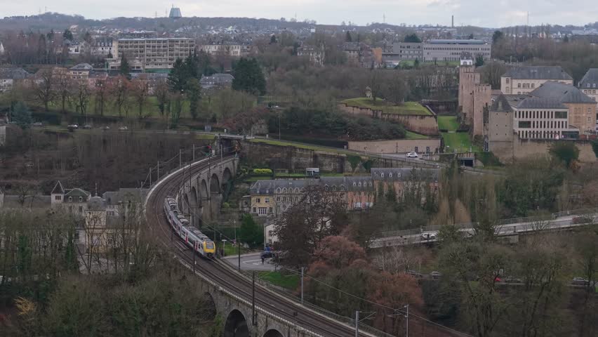 Drone tracking a train passing over a stone bridge in Luxembourg City, capturing urban infrastructure, historic architecture, and dynamic railway movement.