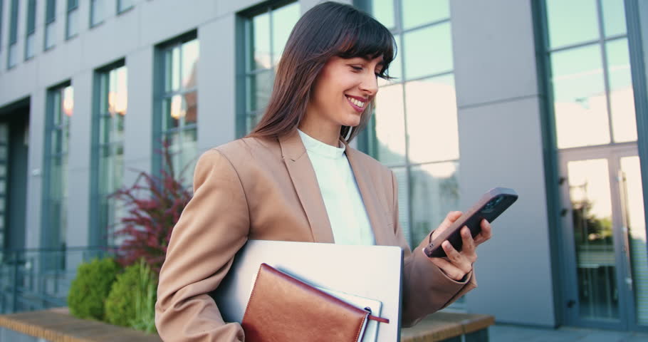 Business woman employee chatting online, texting using a smartphone walking on the street near an office building corporate office typing text messages on mobile phone checking emails successful