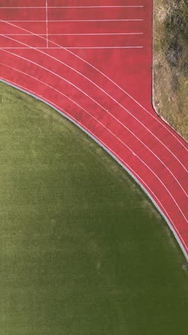 Bird's eye drone footage of a runner in blue clothes jogging along a red athletic track next to a green soccer field. Minimalist geometric composition of an outdoor sports complex.