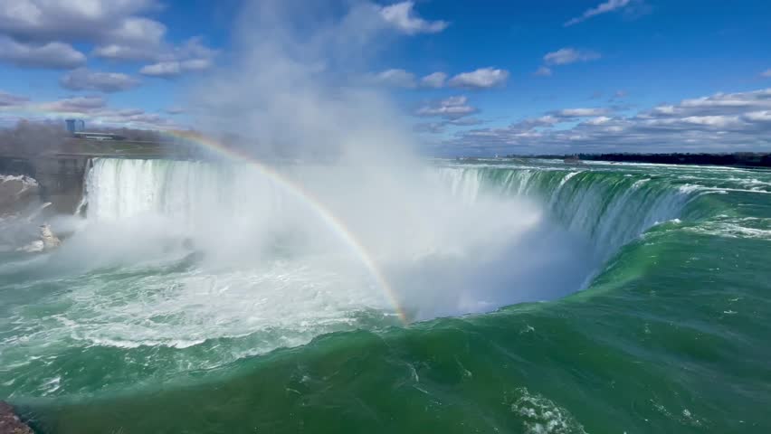 This 4K video captures the Horseshoe Falls of Niagara Falls from the Ontario, Canada shoreline. The visual documents the high-volume water flow over the crest and a rainbow formed by sunlight refracting through the rising mist. The composition illustrates the hydrological power of the Niagara River, documenting North American geography for nature and editorial media.