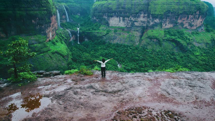 4K Aerial shot of woman standing on cliff with stretched arms and looking at majestic Devkund waterfall in green Sahyadri hills at Maharashtra, India. Drone shot of Devkund waterfall viewpoint at Savalya ghat.