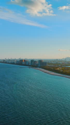 View from the ocean towards a long strip of sandy beach, green vegetation, and distant Miami city buildings. Aerial landscape of sandy beach and coastal city skyline. Vertical video.