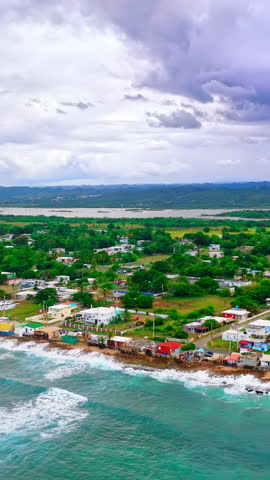 Residential area with colorful houses along a rocky shoreline and green hills in the background. Aerial view of coastal village near turquoise ocean water in Puerto Rico. Vertical video.