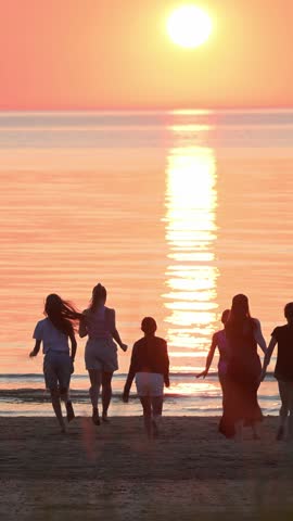 Silhouettes of a group of joyful people running towards the sea and sun on a sandy sea beach in the backlight at sunset. 