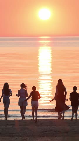 Silhouettes of a group of joyful people running towards the sea and sun on a sandy sea beach in the backlight at sunset. 