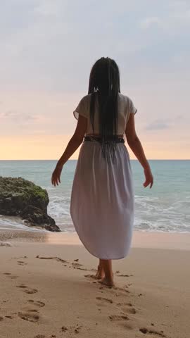 Young Woman with Flowing Dreadlocks Raises Her Arms on Tropical Sunset Beach, Barefoot on Golden Sand. Wearing Light Beige Maxi Skirt and White Cropped Top, She Embodies Freedom, Joy, and Empowerment
