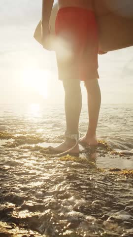 A fit man, male surfer or active guy is stepping into shallow ocean water with a surfboard under his arm, surrounded by gentle waves and glowing reflections. The strong backlight creates a soft