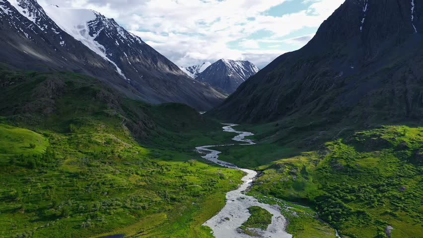 Meandering river flows through a lush green valley between high mountains with snow-capped peaks under a partly cloudy sunny sky. The dramatic landscape combines vibrant greenery, flowing water, and
