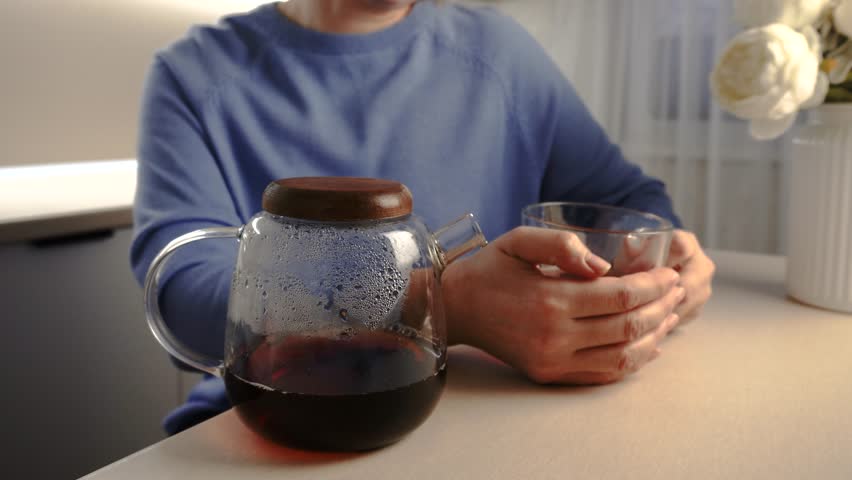 A woman in a blue sweater holds a clear glass cup while sitting near a wooden table. A glass teapot filled with dark liquid sits on the table, showcasing condensation on its surface.