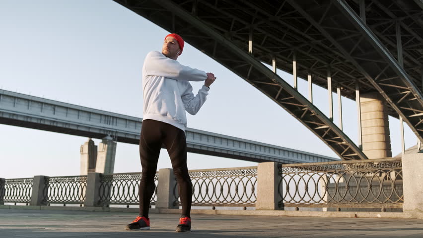 Young athlete wearing a red beanie and white sweatshirt stretching his arm and shoulder before a run near a city bridge. The urban setting and early daylight highlight preparation for an outdoor