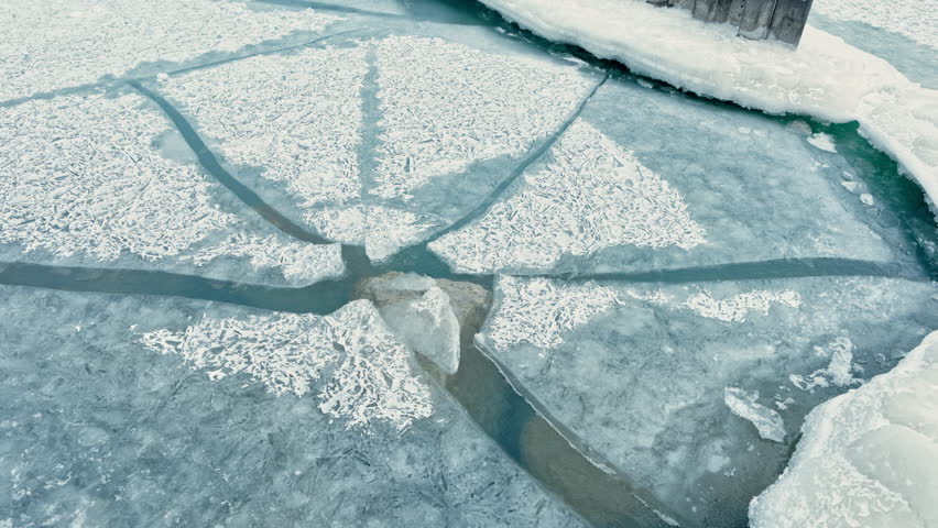 In a rural setting, ice covers a lake creating unique patterns. Cracks and water lines are visible under the frost. It is daytime with natural light brightening the scene.