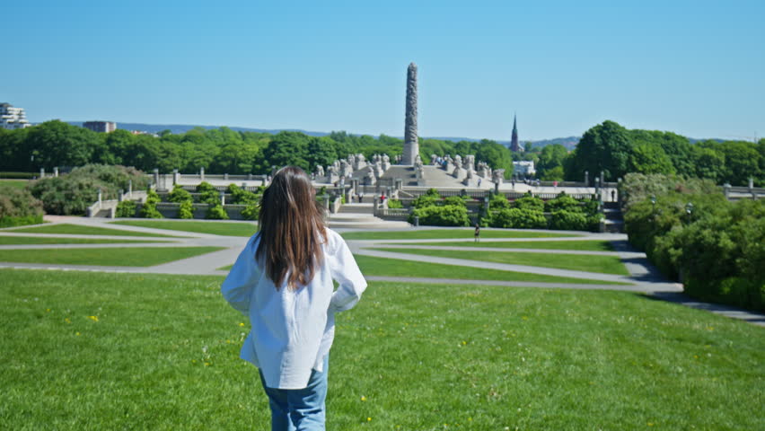 Tourist woman walking across green lawn toward the Monolith sculpture in Vigeland Sculpture Park. Monolith tower, sculpture garden and park landscape during summer in Oslo, Norway.