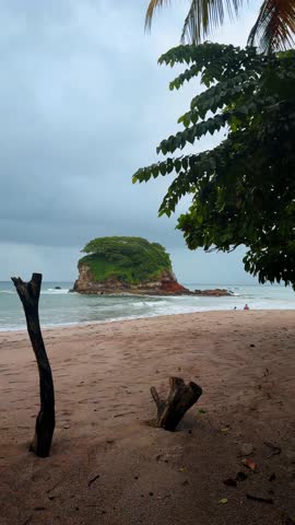 Small rocky island with green vegetation off sandy beach coast. Scenic view of a tropical island near the shoreline under an overcast sky in Costa Rica