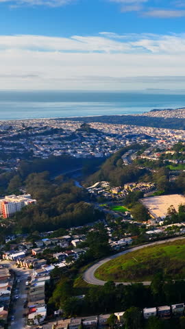 Suburban houses, a winding road, and a lush green valley leading towards the sea under a blue sky. Coastal residential neighborhood near ocean and green valley San Francisco. Vertical video.