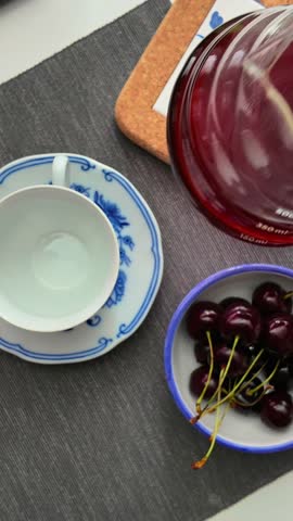 A hand pours filter coffee from a glass teapot into a cup standing on a gray table along with a bowl of cherries