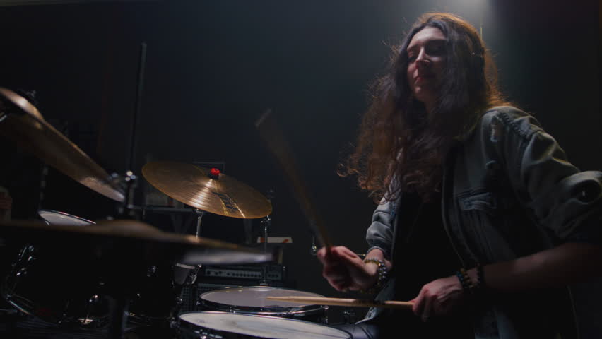 Portrait: female with long curly hair plays the drums. Girl is rehearsing in a studio with drumsticks. Face of a beautiful musician girl, showcasing skill and energy. Underground indie slow motion