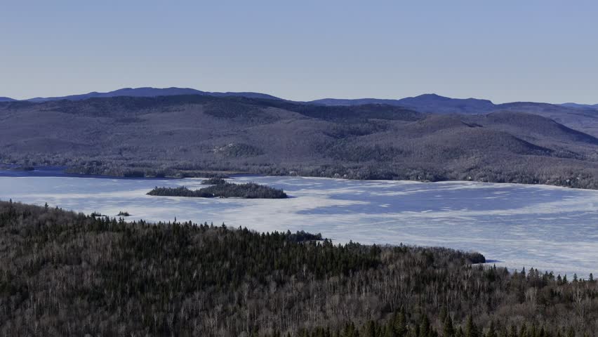Aerial view of frozen lake in Quebec with forest and mountains under blue sky. g.