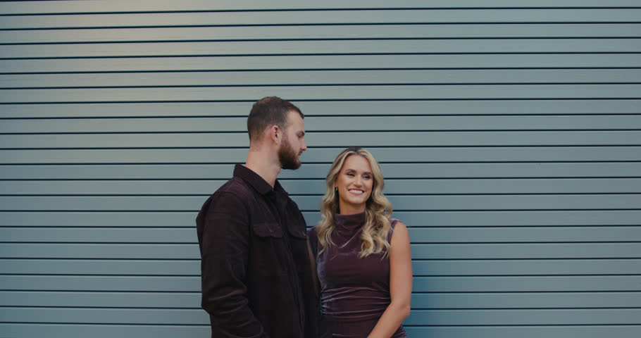 An emotional shot of a loving couple standing against a modern gray wall. The man gently kisses his woman, and she smiles sincerely.