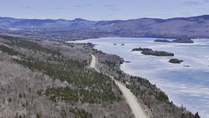 defaultAerial view of winding road along frozen lake in Quebec with forest and mountains under blue sky. g.