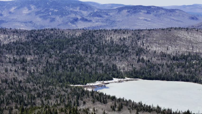Aerial view of frozen lake in Quebec with forest and mountains under blue sky. g.