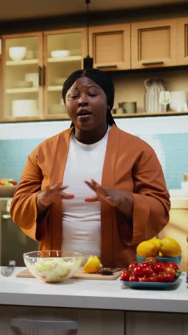 Black female influencer mixing salad ingredients in a bowl, filming gourmet video meal prep at home. Content creator following the cooking process and recording food vlog for audience.