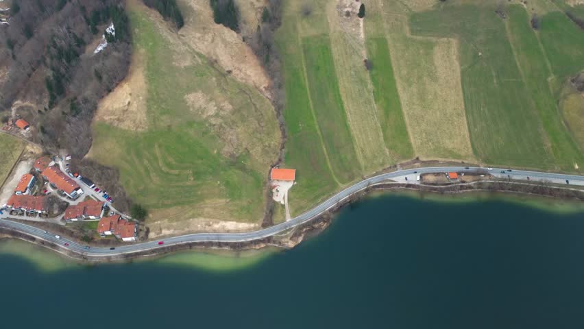 A breathtaking and high-resolution drone aerial view of a picturesque European countryside road winding along a crystal-clear turquoise lake, featuring charming red-roofed houses and lush green agricultural fields under a soft daylight sky, capturing the serene harmony between rural architecture and natural beauty in a professional 4k cinematic panorama