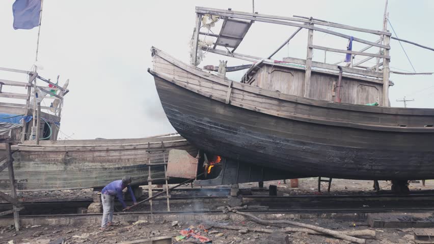 A skilled worker applies fire to the underside of a wooden fishing boat during maintenance in a traditional shipyard. The scene highlights age-old techniques used in boat building and repair, showcasing craftsmanship and maritime industry practices.