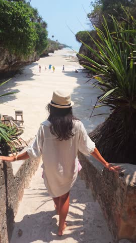 Woman in a straw hat stands at a scenic stone viewpoint overlooking Mtende Beach in Zanzibar. Dramatic coral cliffs frame a secluded white sand cove at low tide.