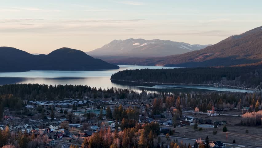 Drone aerial view of a lakeside town surrounded by forest and mountains with calm waters under a pastel sky.