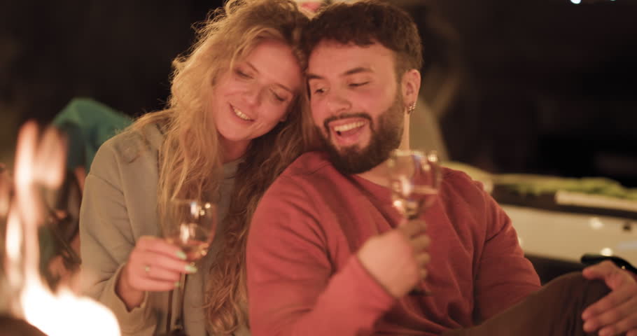 People sit by a campfire in the woods at night. A couple shares wine and smiles together while enjoying a special moment. Friends gather around and laugh under the starry sky.