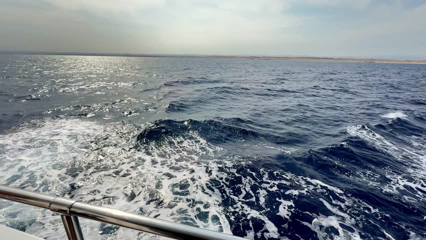 Luxurious yacht glides through Red Sea waves on a breezy spring morning in Egypt under a hazy sun featuring shimmering blue water and silver chrome railings from a wide side deck camera perspective
