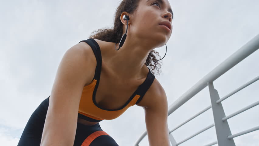 Woman, mark and start running in city with earphones, intense exercise or endurance training outdoor. Low angle, person or runner breathing on bridge with audio tech, begin sprint and cardio workout.