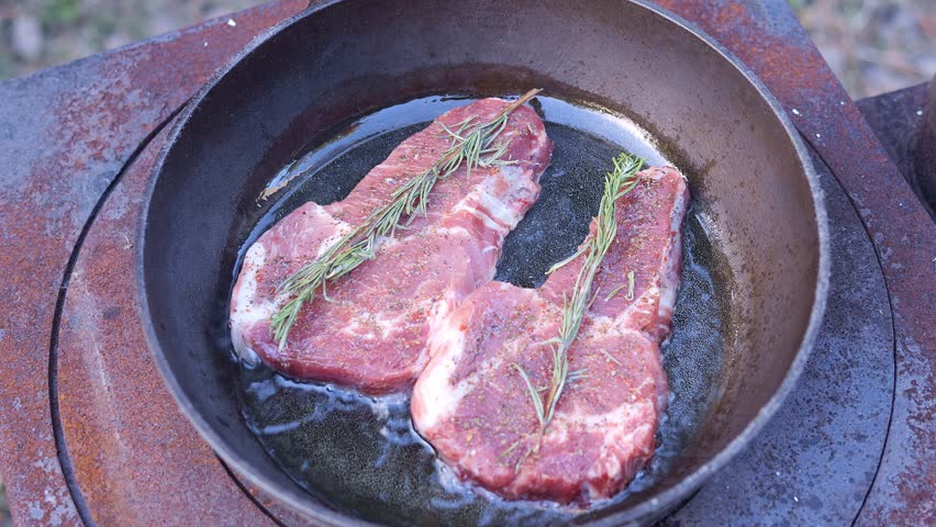 Outdoor cooking frying steak with rosemary on cast iron pan over fire steak sizzling in hot oil steak turning with tongs rustic cooking scene food preparation outdoors camping meal preparation flavor