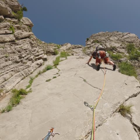 Low belayer view of a lead climber ascending a steep slab route with rope and bolts visible in bright natural sunlight, ideal for climbing and adventure themes