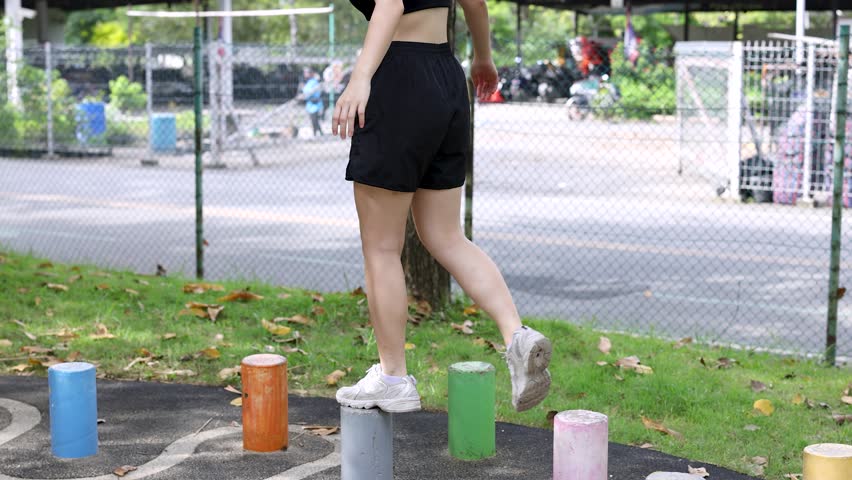 An Asian woman in black sportswear balances on colorful stepping stones in a sunny park