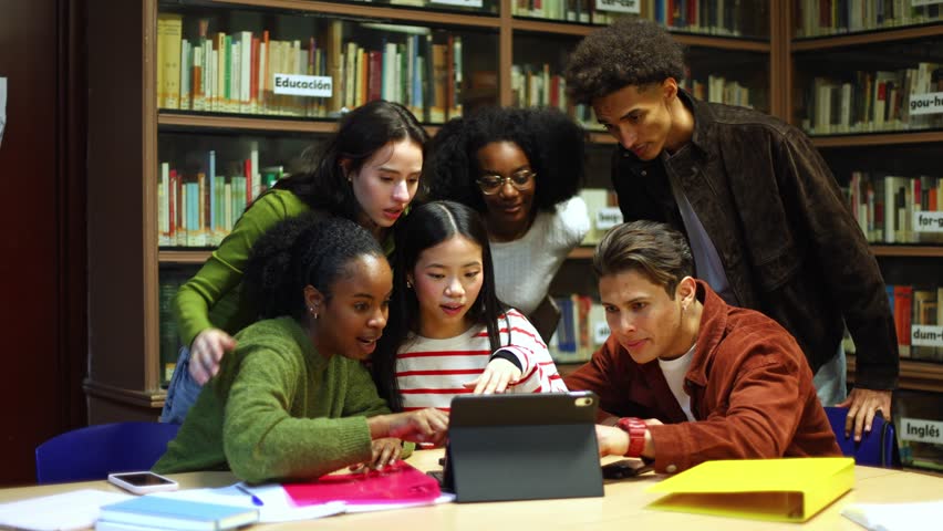 Young multiethnic university students celebrating an achievement while studying together in a library. Happy friends looking at a tablet