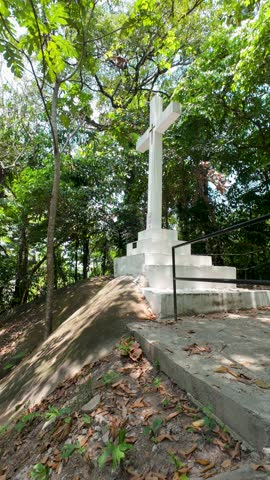 A vertical video of the Stations of the Cross Station 14 set along a serene tropical hillside, featuring a white cross monument surrounded by lush greenery. This video captures a quiet and contemplative atmosphere ideal for themes of faith, reflection, sacrifice, and spirituality. Suitable for Christian content, church presentations, Good Friday observances, Easter storytelling, and meditative visual projects.