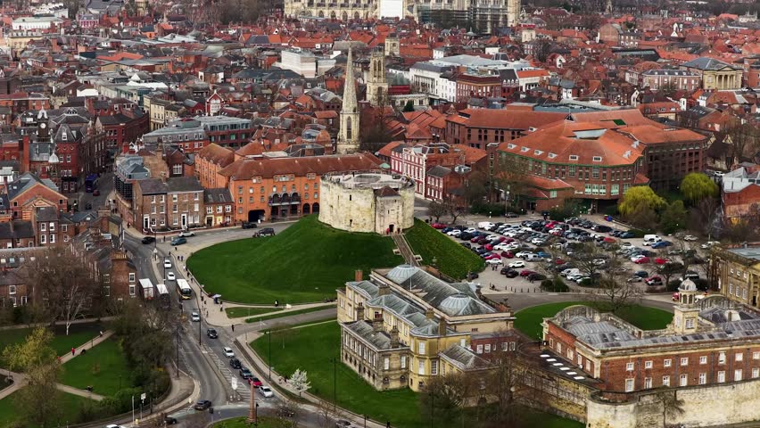 Aerial drone footage of Clifford Tower in the city of York in Yorkshire UK showing circular structure grass mound surrounding roads and car park captured directly from above in the spring time