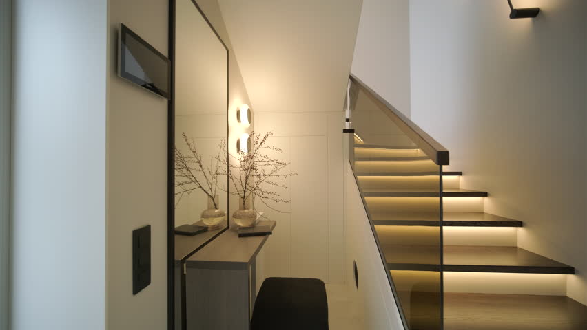 Modern interior design of a wooden staircase with integrated warm LED strip lighting, a glass balustrade, and a large mirror in a minimalist hallway.