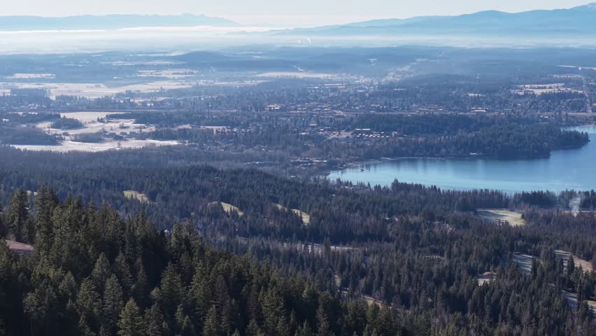 Aerial view of a dense pine forest and shimmering lake surrounded by mountains near Whitefish, Montana.