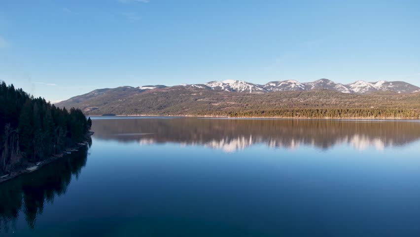 Aerial panorama of a calm lake reflecting snow-capped mountains and clear blue sky near Whitefish, Montana.