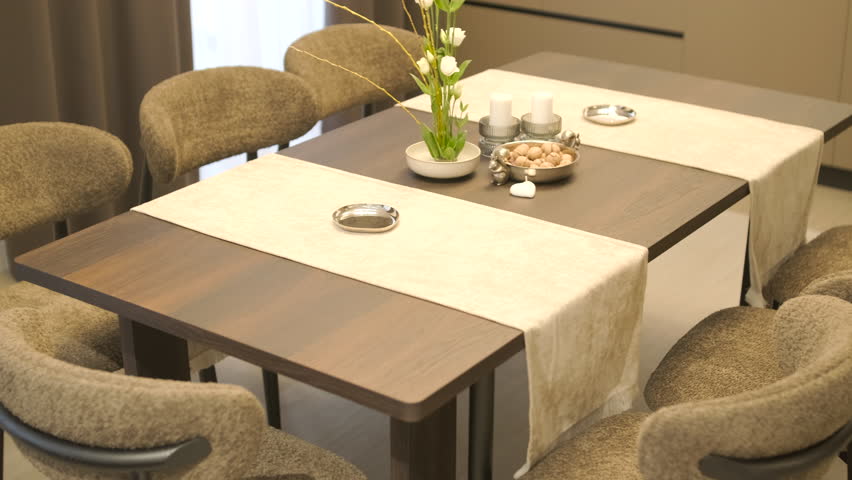 Elegant dining table setting in a modern interior, featuring white burning candles, a floral arrangement with white flowers, and decorative bowls on a dark wooden table with beige runners.