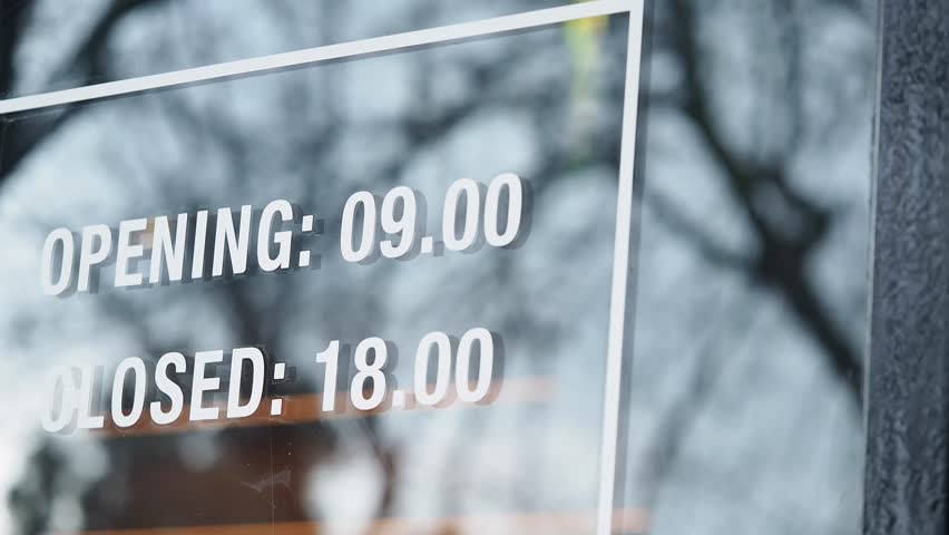 Opening and closing hours sign on a glass door, showcasing a reflection of trees, business operating hours displayed clearly