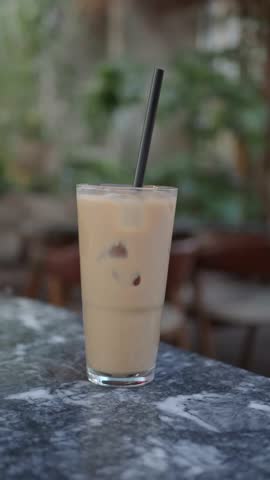 Close-up shot of a refreshing iced coffee beverage with ice cubes and a black straw on a marble table, as a hand enters the frame and stirs it gently